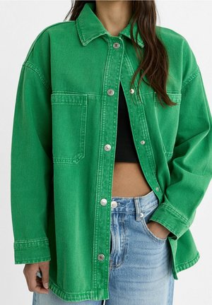 Personne portant une veste en jean verte oversize à boutons, un crop top noir et un jean bleu clair avec une main dans la poche.