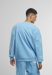 Felpa azzurra chiara in materiale morbido, con vestibilità comoda, spalle scese e piping bianco sui lati. La vista dal retro mostra dettagli minimi.