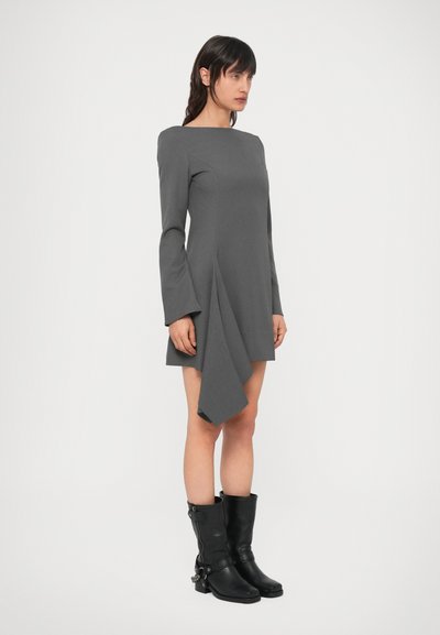 Szürke, hosszú ujjú ruha hajónyakú kivágással, aszimmetrikus szegéllyel és bővülő ujjakkal. Fekete térdig érő csizmával párosítva.