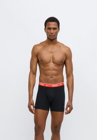 Fekete boxeralsó piros Nike-logós derékrésszel, testhez simuló kialakítással és sima anyagú textúrával. Modell egyszínű háttér előtt pózol.