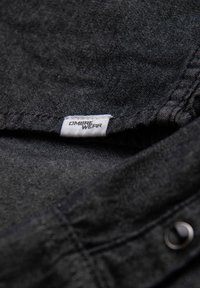 Vêtement en tissu noir avec surface texturée. Comprend une étiquette de logo portant l'inscription "OMBRE WEAR" et un détail de bouton. Gros plan.