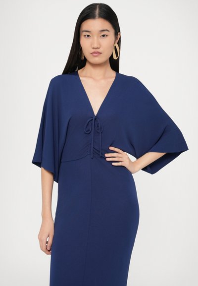 Φόρεμα σε ναυτικό μπλε με βαθύ V-ντεκολτέ, ζώνη στη μέση με δέσιμο και φαρδιά μανίκια. Κατασκευασμένο από απαλό ύφασμα με εφαρμοστή σιλουέτα.