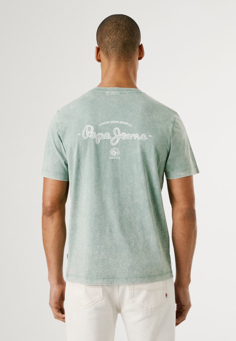 T-shirt en coton vert avec un col rond, des manches courtes et une finition délavée. Le dos affiche un logo imprimé en blanc et des détails textuels.