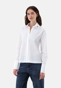 Camicetta bianca a maniche lunghe con colletto strutturato, leggero lucido e dettagli decorativi sul colletto. Abbinata a jeans.