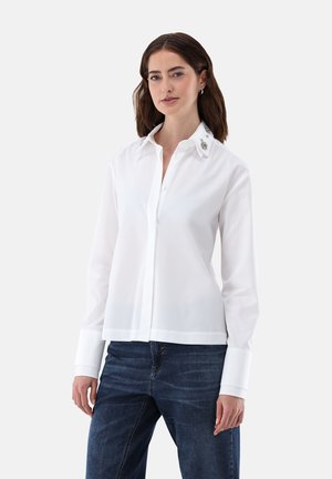 Camicetta bianca a maniche lunghe con colletto strutturato, leggero lucido e dettagli decorativi sul colletto. Abbinata a jeans.