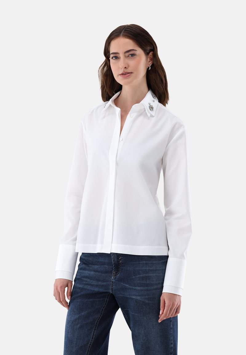 Camicetta bianca a maniche lunghe con colletto strutturato, leggero lucido e dettagli decorativi sul colletto. Abbinata a jeans.