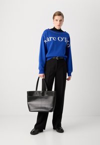 Zils džemperis ar baltu uzrakstu, melni bikses un melna ādas rokas soma. Apģērbs ir brīvs piegriezumā un ar elegantu siluetu.