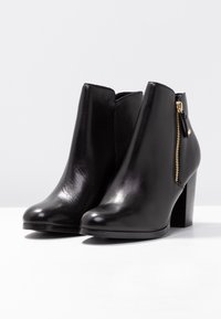 Botas de piel negra hasta el tobillo con punta afilada, detalle de cremallera lateral y tacón grueso. Textura suave y diseño elegante.
