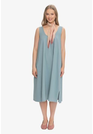 Kobieta nosząca luźną, niebieską sukienkę midi bez rękawów, różowy szal zawiązany wokół szyi i różowe płaskie buty, stoi i się uśmiecha.