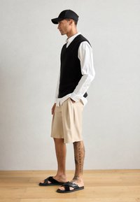 Svart ribbestrikket vest over hvit stripete skjorte, beige plissert shorts og svarte sandaler; vist mot en nøytral bakgrunn.