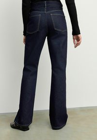 Des jeans évasés en denim foncé présentent une taille haute, des poches arrière proéminentes, des coutures contrastantes et une texture lisse. Chaussures noires à talon bas.