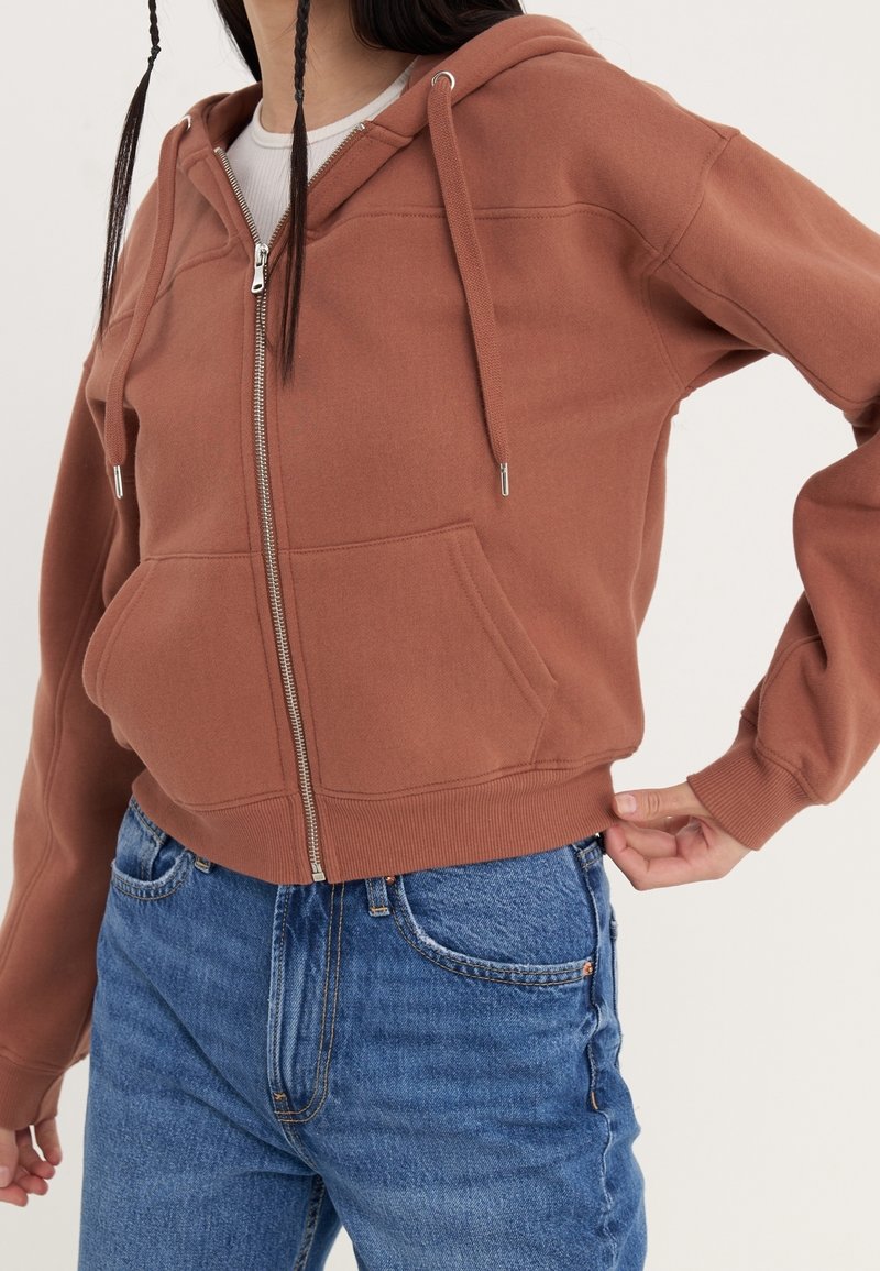 Sudadera corta marrón con cremallera frontal, bolsillo delantero, capucha con cordón y puños acanalados. Combinada con vaqueros azules de talle alto.