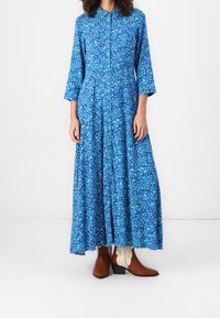 Lang, knap-op kjole i blå med blomsterdesign. Har trekvartlange ærmer og en fuld, svajende nederdel. Bæres med brune ankelstøvler.