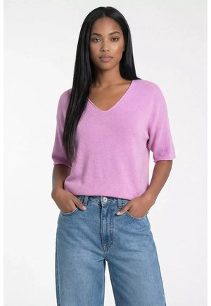 Kobieta z długimi, prostymi czarnymi włosami, ubrana w jasnofioletowy sweter z krótkim rękawem i dekoltem w serek oraz wysokim stanem niebieskie dżinsy, z rękami w kieszeniach.