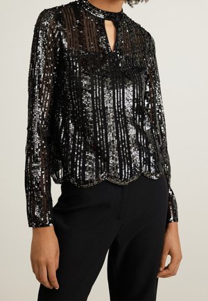 Sort transparent langærmet bluse med lodrette sølvpalietter, nøglehulshalsudskæring og bølget kant, kombineret med sorte bukser, båret af en person.