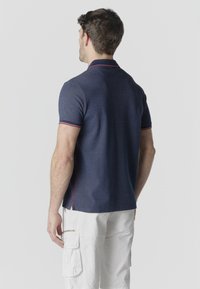 Uomo di spalle che indossa una polo a maniche corte blu navy con rifiniture rosse e pantaloncini cargo bianchi su uno sfondo chiaro e uniforme.