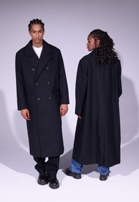 Manteau noir croisé avec de grands boutons et des poches latérales, présentant une coupe longue et une texture lisse, porté sur un pantalon sombre.