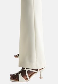 Sandali bianchi con tacco alto, caratterizzati da un design a strisce e punta affilata, abbinati a pantaloni tailored color crema, che mostrano linee pulite e una texture morbida.