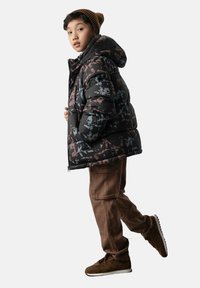 Dziecko w wzorzystej czarnej kurtce puchowej, brązowych spodniach cargo, brązowych butach i brązowej wełnianej czapce, stojące z delikatnym odchyleniem do tyłu.