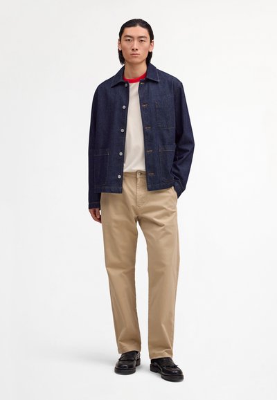 Νέος άνδρας στέκεται φορώντας σκουρό μπλε τζιν μπουφάν, λευκό και κόκκινο μπλουζάκι, μπεζ παντελόνι και μαύρα παπούτσια σε απλό λευκό φόντο.