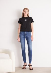 Zwarte katoenen t-shirt met een logo aan de voorkant, gecombineerd met blauwe skinny jeans. Model draagt zwarte hoge sandalen. Eenvoudige, casual look.