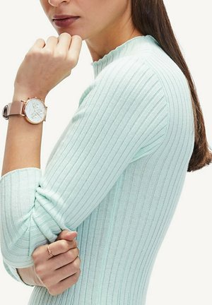 Minzgrünes, geripptes Langarmoberteil mit einem gewellten Ausschnitt. Tragen mit einer runden Uhr mit goldenen Akzenten, die ein weißes Ziffernblatt und ein helles Armband hat.