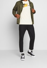 Person som visar en outfit med olivgrön jacka, vit Nike-t-shirt med orange logga, svarta byxor, vita strumpor och svarta Nike-sneakers.