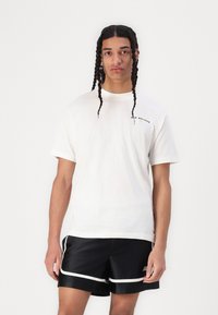 Fehér pamut póló kerek nyakkal, fekete és sárga "New Balance" logóval. Fekete selyemsort visel fehér csíkokkal.