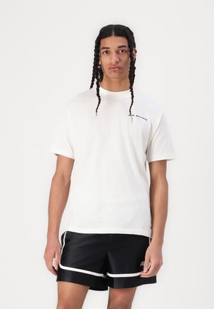 Weißes Baumwoll-T-Shirt mit Rundhalsausschnitt, versehen mit dem "New Balance"-Logo in Schwarz und Gelb. Kombiniert mit schwarzen Satin-Shorts mit weißen Streifen.