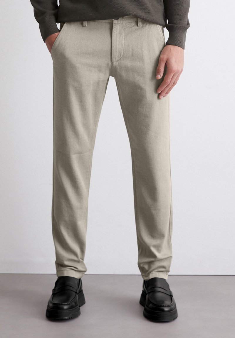 Beige Hose mit geradem Schnitt und subtiler Textur, ausgestattet mit Fronttaschen, getragen mit schwarzen Schuhen.