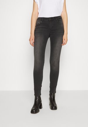 Vrouw draagt zwarte skinny jeans en zwarte enkellaarsjes met hak, staand tegen een effen lichte achtergrond.
