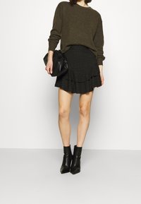 Pull en tricot vert olive, jupe mini noire à volants avec détails œillets, et bottines noires, tenant un sac à main noir.