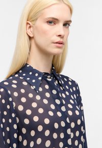 Marineblaue transparente Bluse mit großen crémefarbenen Pünktchen, klassischem Kragen, Knopfleiste und langen Ärmeln. Leichtes Material mit glatter Textur.