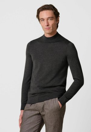 Mann mit heller Haut und braunen Haaren, der einen dunkelgrauen Rollkragenpullover und hellbraune Hosen trägt, stehend mit einer Hand in der Tasche.