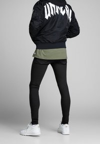 Chaqueta bomber negra con grandes letras blancas en la espalda, superpuesta a una camiseta verde oliva y combinada con jeans negros ajustados y zapatillas blancas.