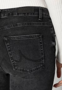 Jeans de denim preto com um acabamento texturizado, apresentando um bolso traseiro com um logo costurado e um patch em couro preto na cintura.