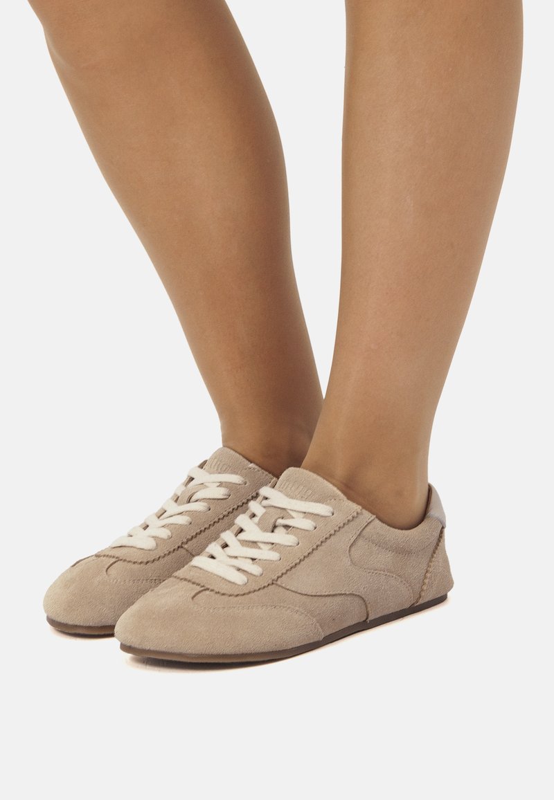 Zapatillas deportivas de ante beige con cordones blancos, que presentan un perfil bajo, puntera redondeada y detalles sutiles de costuras. Textura suave y diseño minimalista.