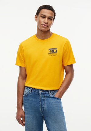 Νεαρός άνδρας με κίτρινη κοντομάνικη μπλούζα με μικρό λογότυπο στο στήθος, φορά μπλε τζιν, στέκεται με το ένα χέρι στην τσέπη μπροστά σε απλό φόντο.