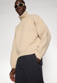 Beige geribbelde gebreide trui met een hoge kraag en verlaagde schouders, gecombineerd met zwarte broek met een ritszakdetail.