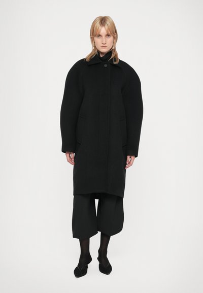 Schwarzer Wollmantel mit übergroßen Schultern, einem Knopfverschluss, seitlichen Taschen und geradem Schnitt, kombiniert mit schwarzen weit geschnittenen Hosen und Schuhen.