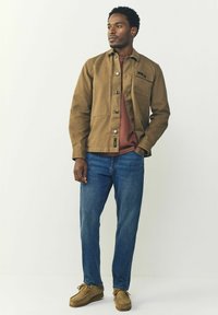 Chaqueta de lienzo marrón con cierre de botones, bolsillo en el pecho, llevada sobre una camisa de color óxido, combinada con jeans azules y zapatos de ante color beige.