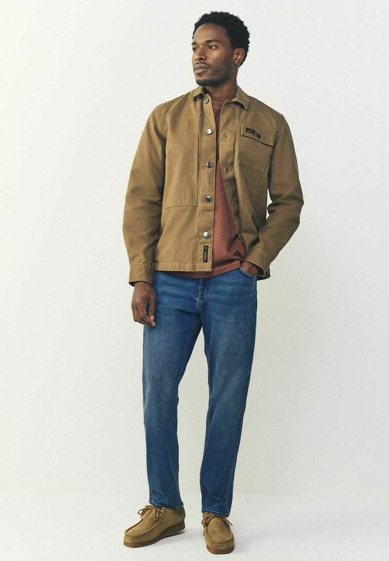 Chaqueta de lienzo marrón con cierre de botones, bolsillo en el pecho, llevada sobre una camisa de color óxido, combinada con jeans azules y zapatos de ante color beige.