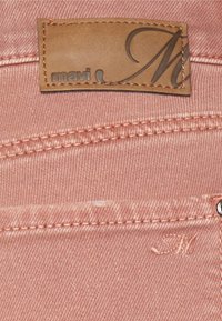Tela de denim rosa con bordes cosidos, que presenta una etiqueta de cuero marrón grabada con "mavi" y un acento de "M".