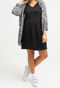 Donna che indossa un vestito nero fino al ginocchio, un cardigan grigio lavorato a maglia e sneakers bianche con calzini bianchi, in piedi con le gambe incrociate.