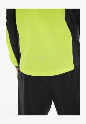 Leichte Jacke in leuchtendem Gelb mit schwarzen Akzenten. Hat ein Herzschlag-Muster, das nahe dem Saum aufgedruckt ist. Glatte Textur, sportliches Design.