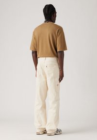 Camiseta de algodón marrón y pantalones de pierna ancha color blanco sucio con bolsillos traseros, que presentan un acento de logo rojo. Hombre de pie con la espalda girada.
