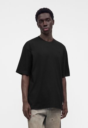 Jeune homme aux cheveux courts et bouclés porte un t-shirt noir ample et un jean clair usé sur un fond uni clair.