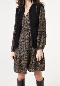 Gilet noir texturé sur une robe fleurie sombre à motifs avec des manches longues. Présente une taille froncée et des poches. Des bottes noires jusqu'aux genoux complètent le look.
