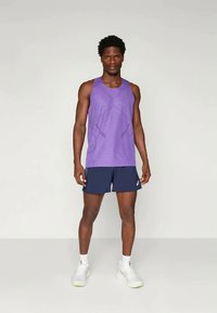 Débardeur de sport violet avec un motif texturé, associé à un short bleu marine. Le modèle porte des chaussures de sport blanches et des chaussettes blanches.