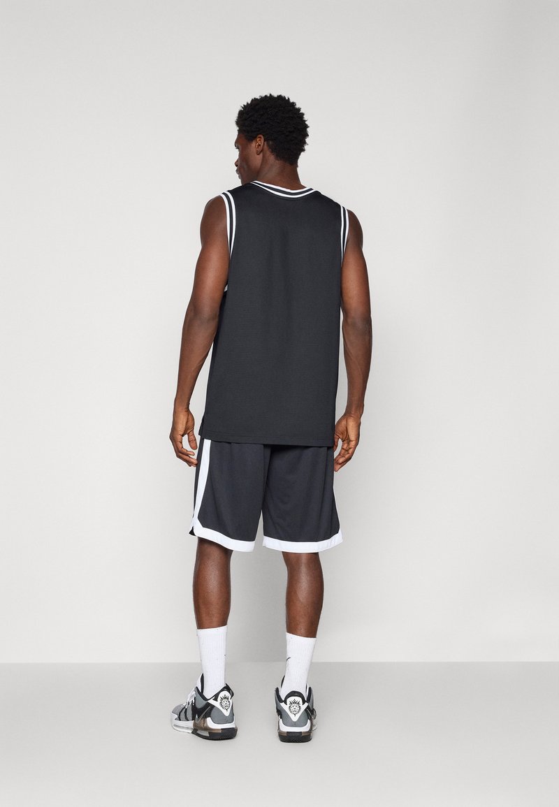 Jersey deportivo sin mangas negro con ribetes blancos, combinado con pantalones cortos de baloncesto negros con acentos blancos y zapatillas deportivas negras.